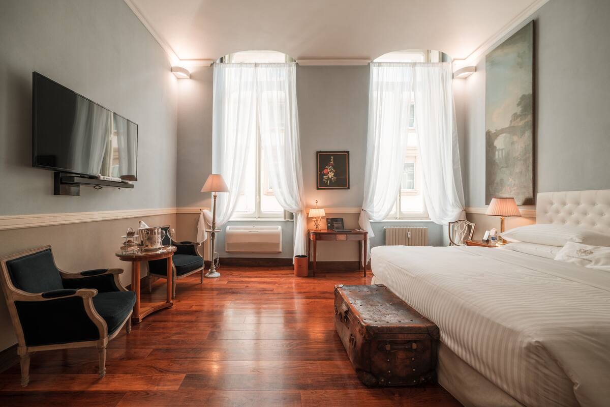 A hotel room with a bed, two chairs, a small table, a lamp, a television, and a painting on the wall.