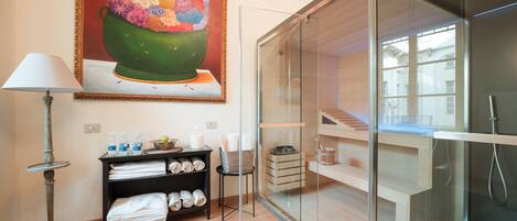 A modern bathroom with a glass shower enclosure, a wooden floor, a towel rack, and a painting of a colorful floral arrangement.