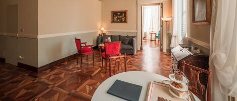Habitación de hotel con piso de parquet de madera, un sofá gris, sillas rojas, una mesa blanca con un libro y un área de comedor con mesa y sillas.