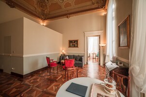 Habitación de hotel con piso de parquet de madera, un sofá gris, sillas rojas, una mesa blanca con un libro y un área de comedor con mesa y sillas.
