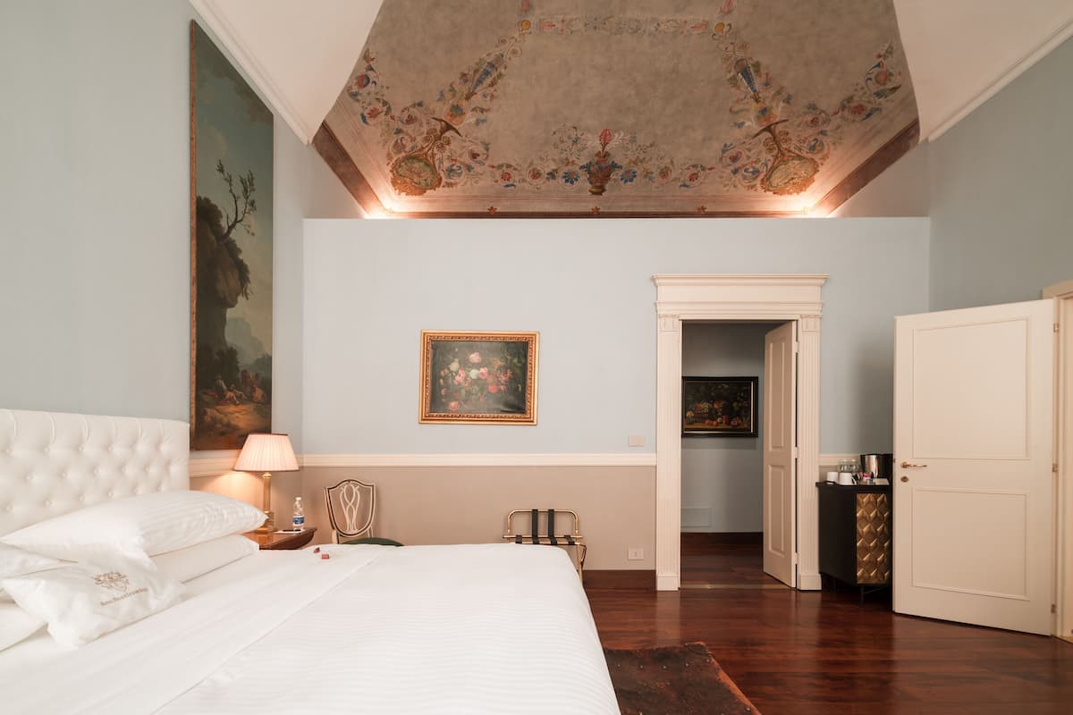 A bedroom with a large bed, a nightstand, a lamp, a painting on the wall, a door leading to another room, and a ceiling with floral patterns.