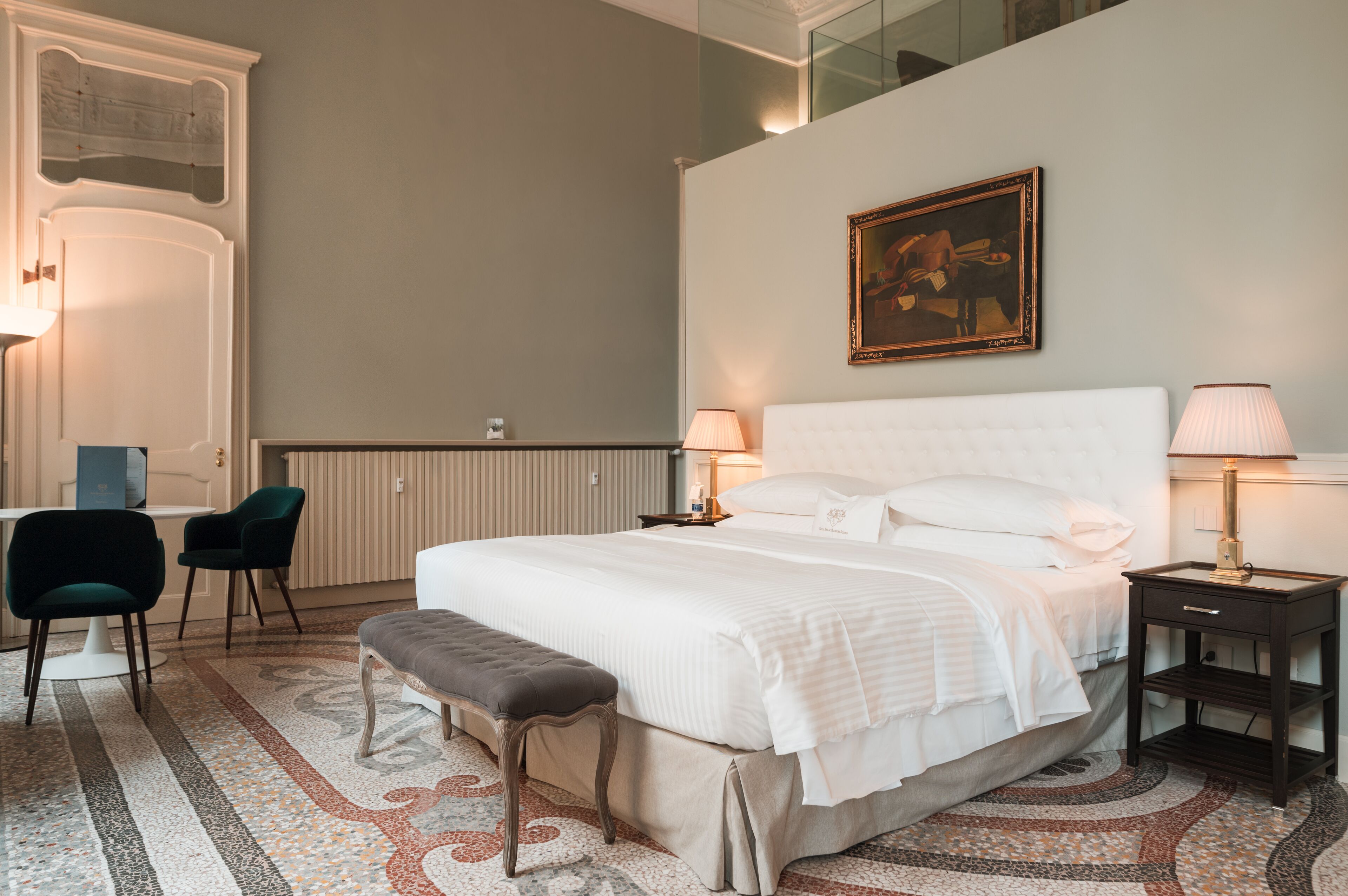 Une chambre d’hôtel avec un grand lit, deux tables de chevet, un banc, un bureau avec des chaises et un tableau au mur.