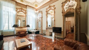 A spacious living room with a large brown leather sofa, a wooden coffee table, and a TV. The room features ornate mirrors, a fireplace, and a chandelier. - Royal Palace Luxury Suites & Hotel (Turin)