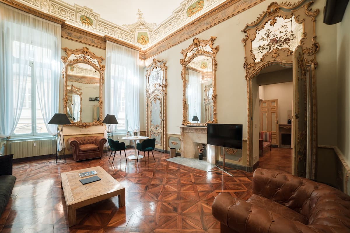 A spacious living room with a large brown leather sofa, a wooden coffee table, and a TV. The room features ornate mirrors, a fireplace, and a chandelier.