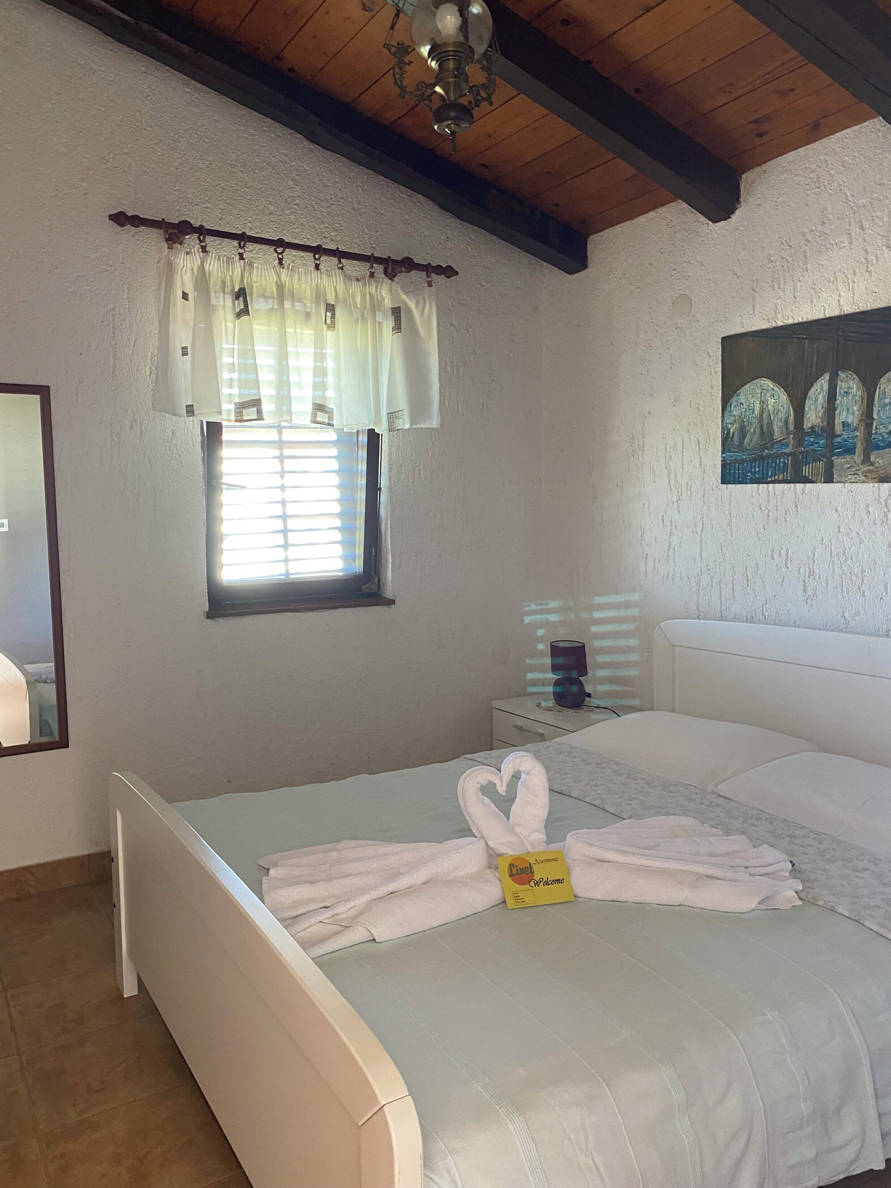 A bedroom with a bed, a window with blinds, a painting on the wall, and a chandelier.