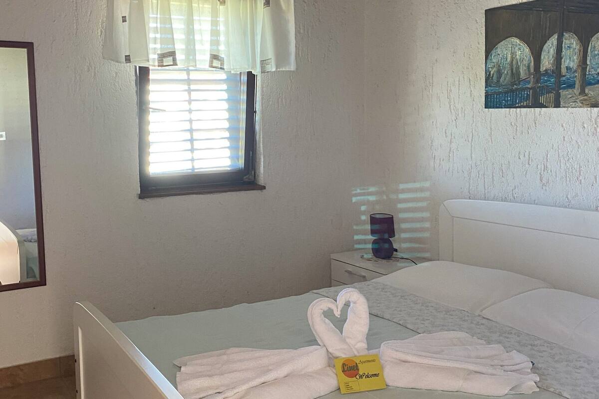 A bedroom with a bed, a window with blinds, a painting on the wall, and a chandelier.