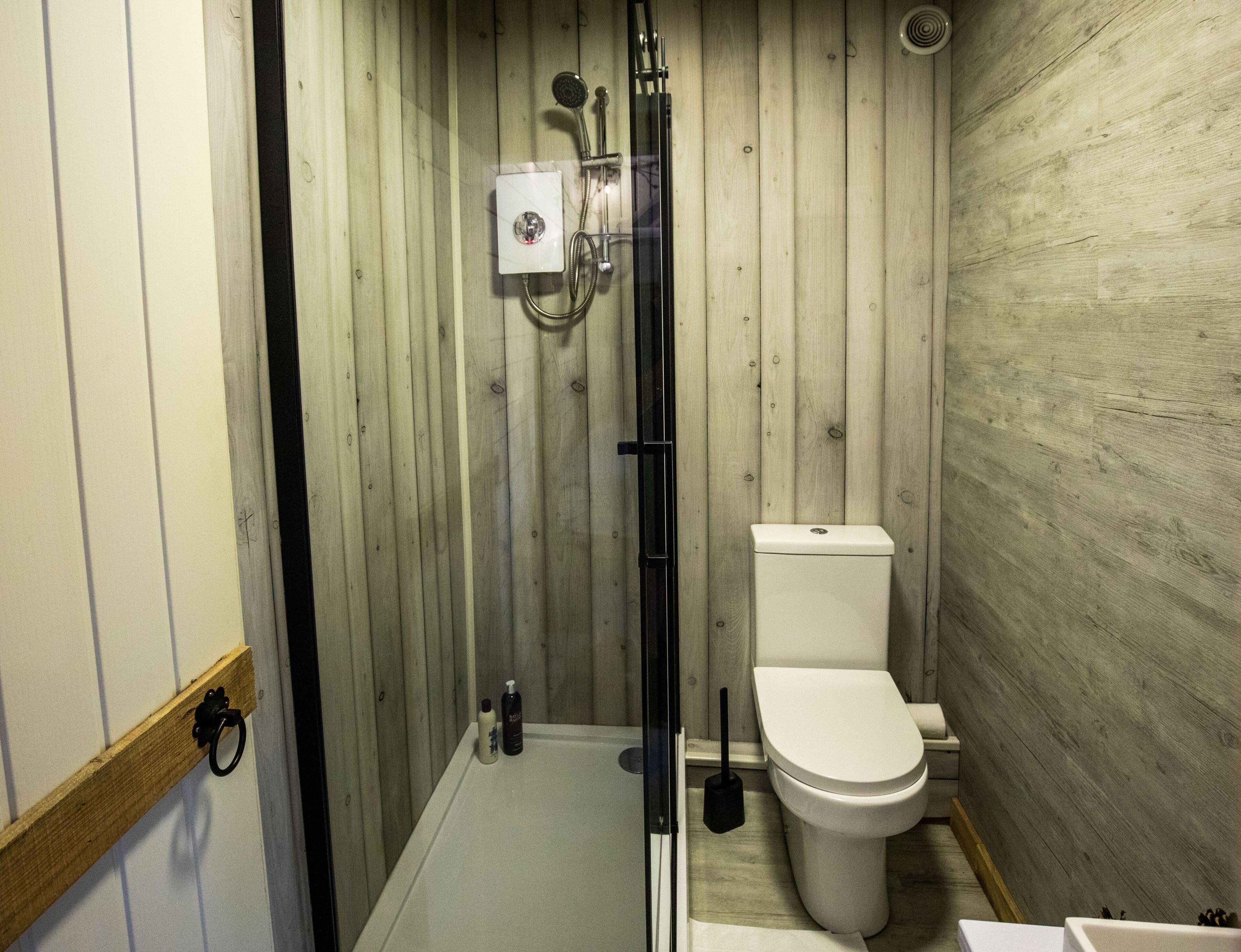 A small, compact bathroom with a wooden wall, a white toilet, a showerhead, and a handheld shower attachment.