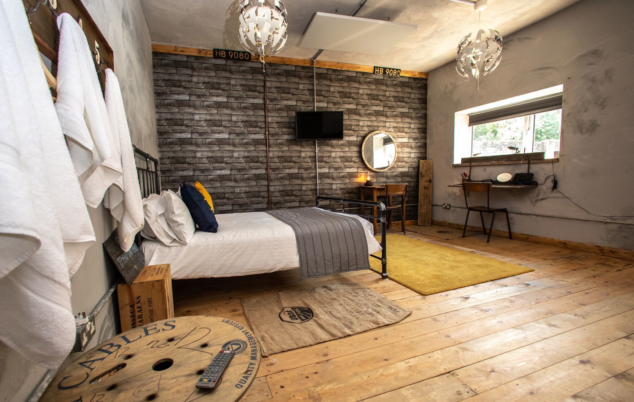 Un dormitorio con cama, suelo de madera, pared de ladrillo, ventana, escritorio y silla.