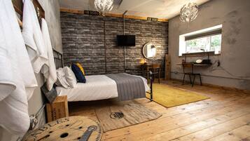 Un dormitorio con cama, suelo de madera, pared de ladrillo, ventana, escritorio y silla.