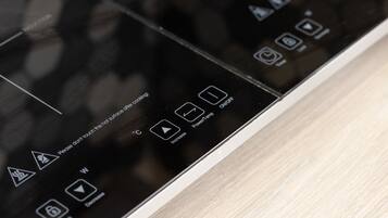 Primer plano de un panel de control negro con varios iconos y texto, incluyendo una advertencia sobre superficies calientes y opciones para aumentar o disminuir la temperatura.