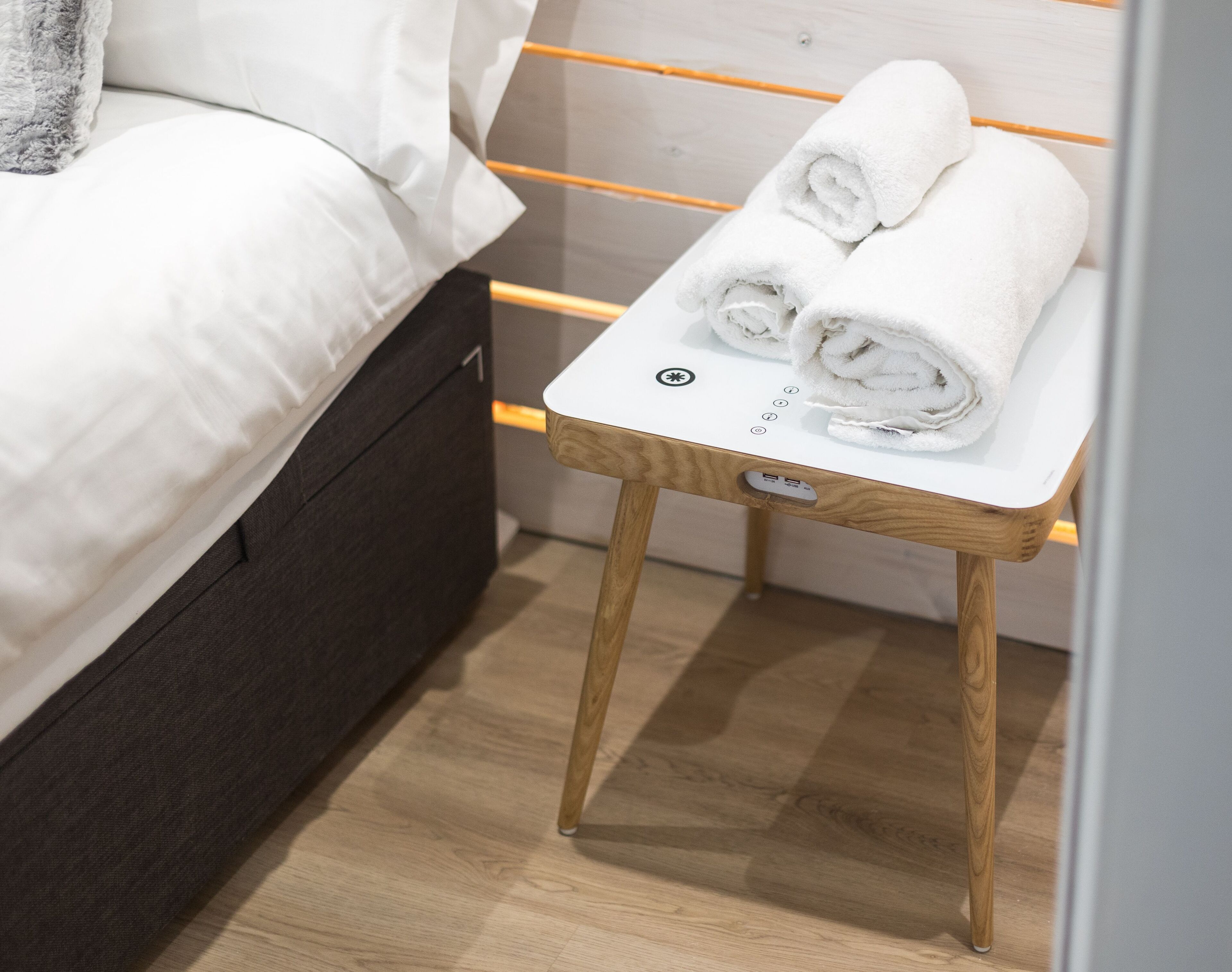 A neatly made bed with white linens and a wooden nightstand with a built-in heater.