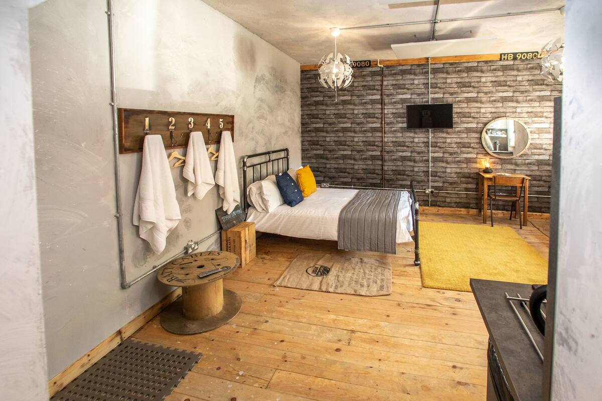 A bedroom with a bed, wooden floor, a mirror, a small table, and a hanging towel rack.