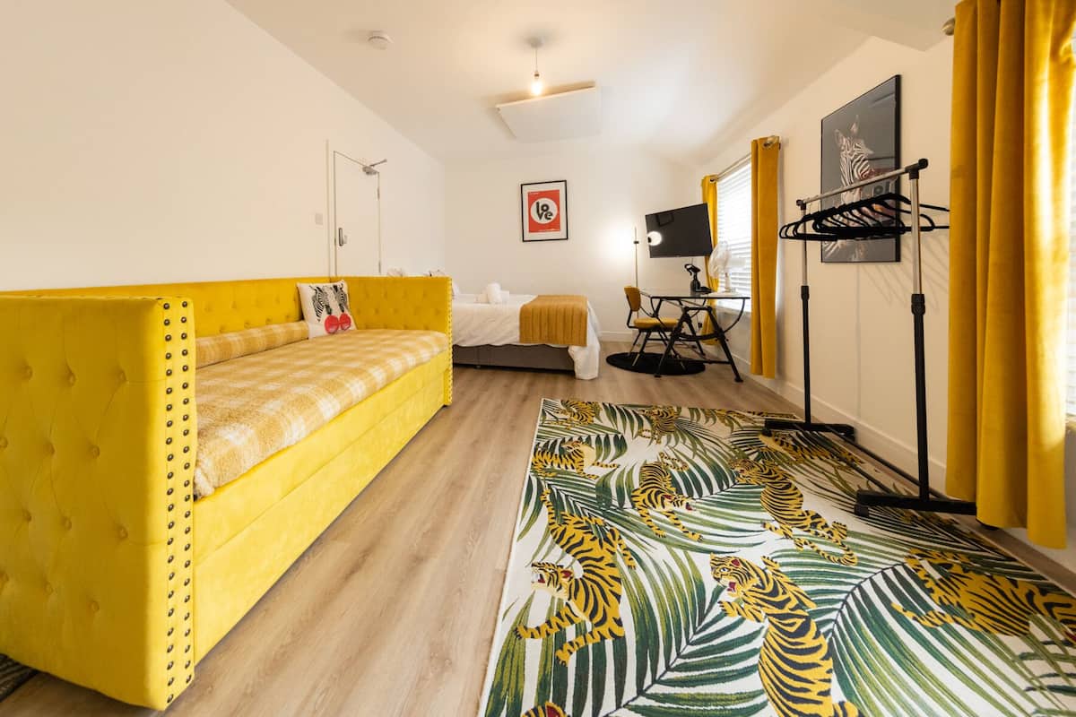 A room with a yellow sofa, a bed, a small table with a chair, a TV, and a zebra picture on the wall.