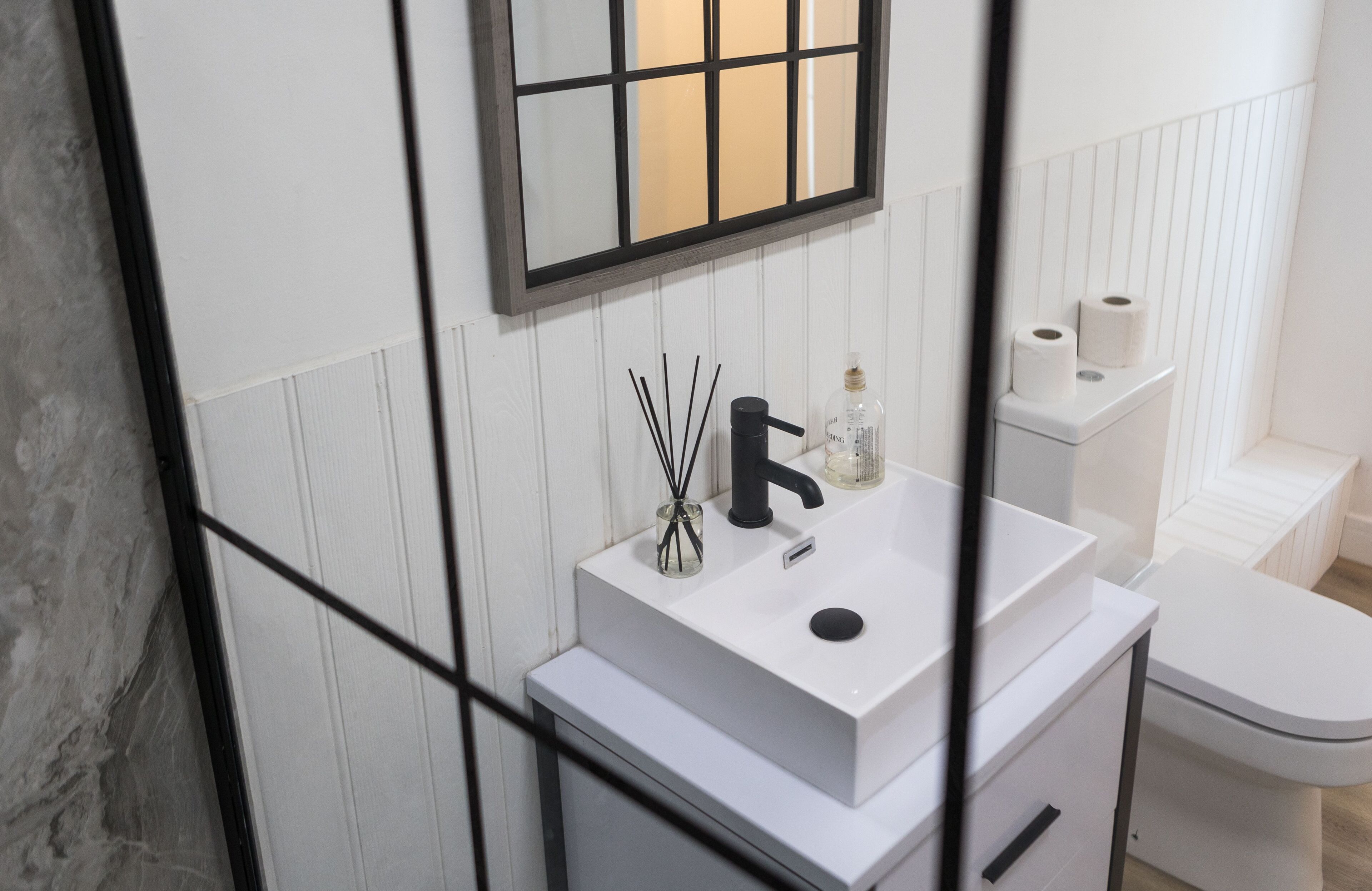 A modern bathroom with a white sink, black faucet, and a mirror above it. There are two rolls of toilet paper on a white shelf.