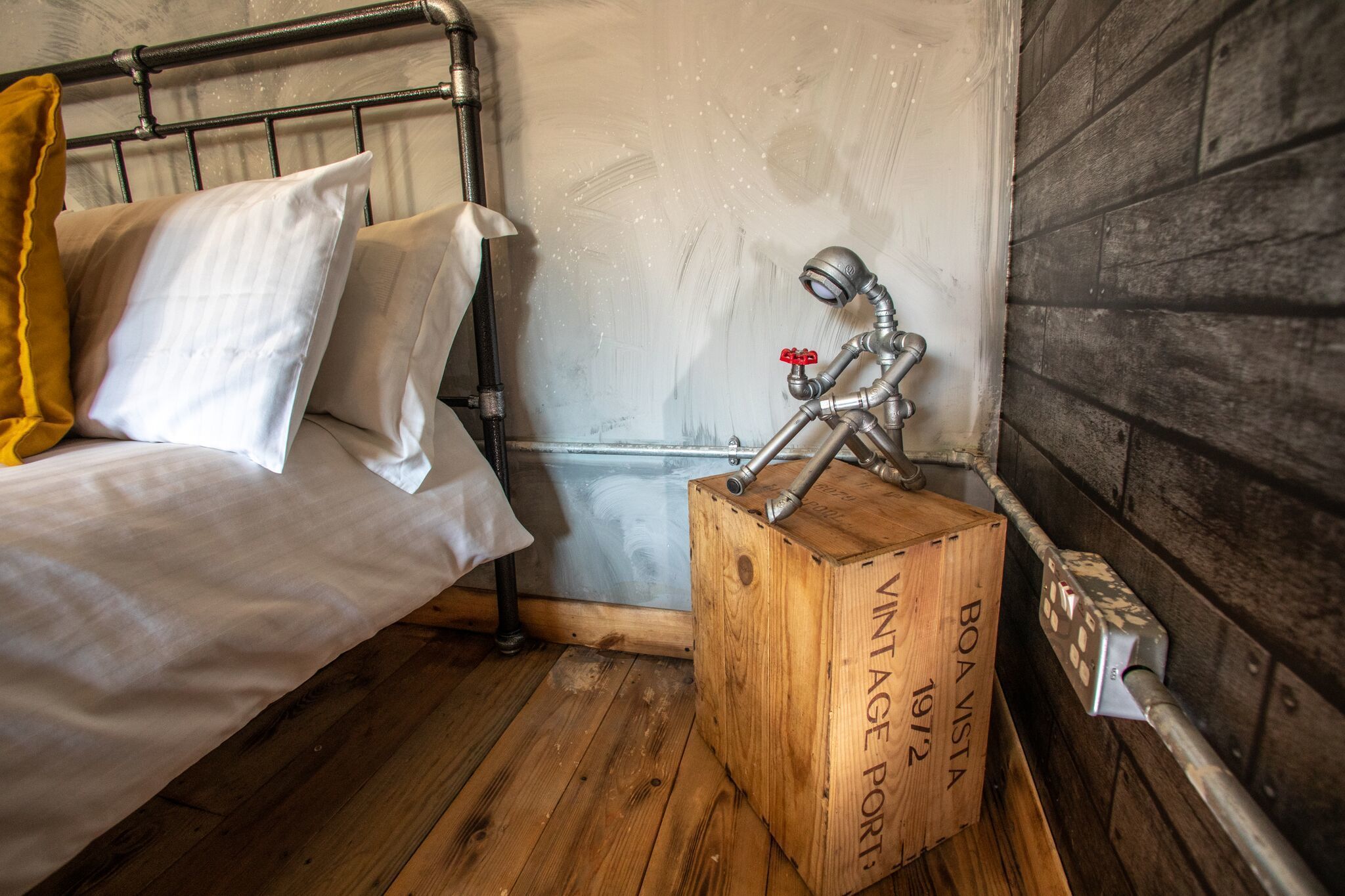 Una cama con sábanas blancas y una almohada amarilla, una caja de madera con la etiqueta "BOA VINTAGE PORT" y una lámpara metálica en el suelo.