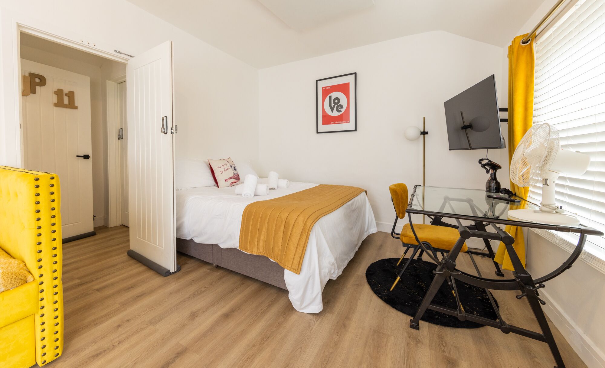 A hotel room with a bed, a desk with a chair, a window with blinds, and a door with a number.