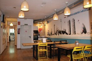 A restaurant with wooden tables and yellow chairs, a mural on the wall, and hanging pendant lights. - Mediterranean Hostel (Barcelona)