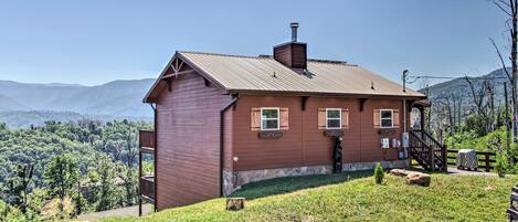 A wooden cabin with a stone foundation, a chimney, and a small porch. It is situated on a grassy hill with a view of a forested mountain range.