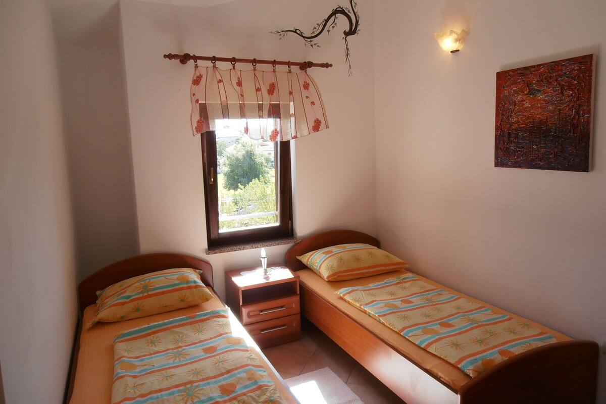 A small, well-lit bedroom with two single beds, a nightstand, a window with a floral curtain, and a wall-mounted light fixture.