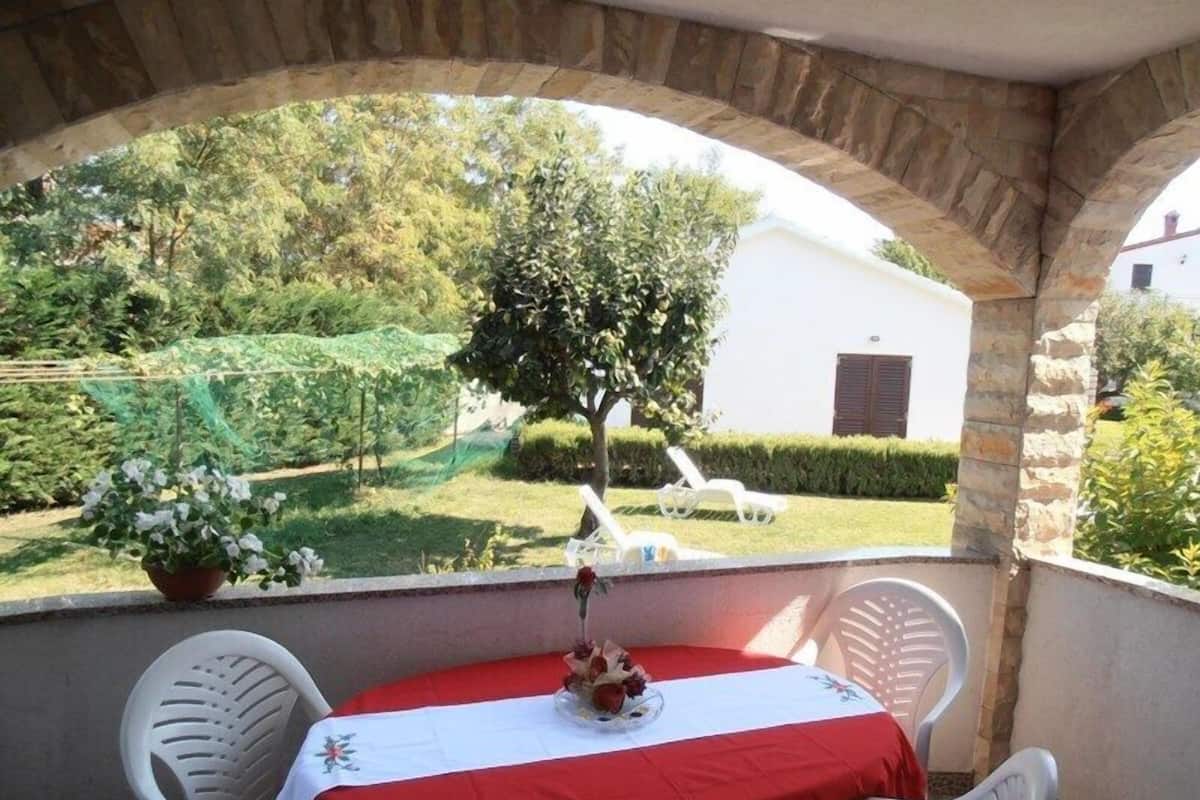 A covered patio with a table set for two, overlooking a garden with a tree and a white building in the background.
