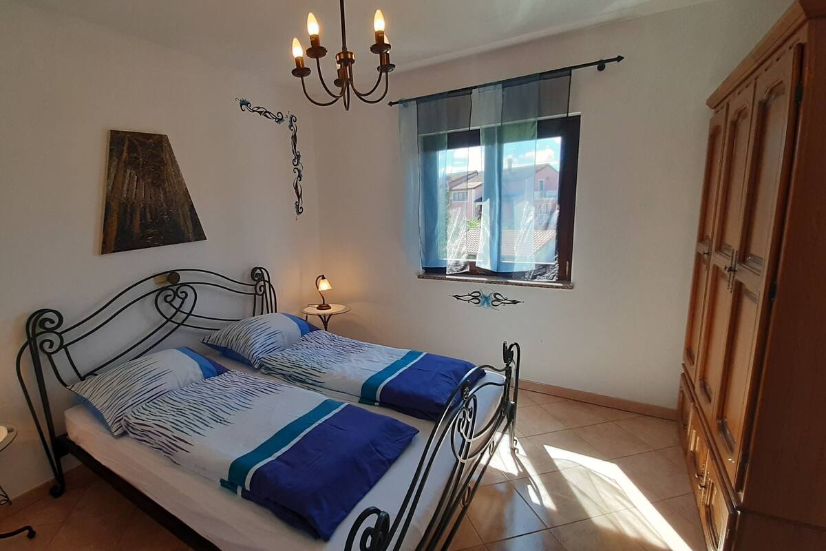 A bedroom with a bed, bedside lamps, a window with curtains, a chandelier, and a wardrobe.