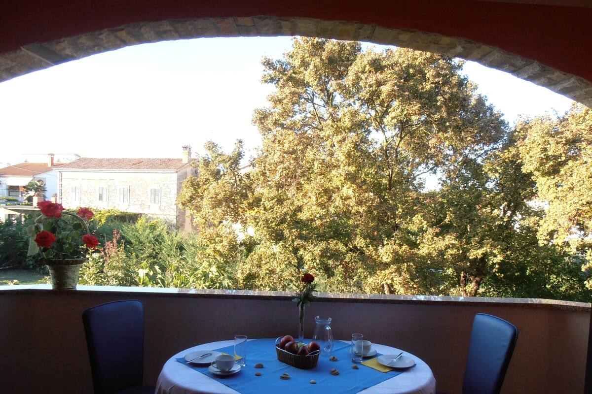 An outdoor dining area with a table set for two, a view of a tree and buildings, and a basket of fruit.