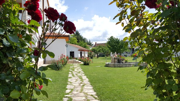 Un sendero en un jardín que lleva a un edificio blanco con techo de tejas rojas, rodeado de abundante follaje y rosales rojos.