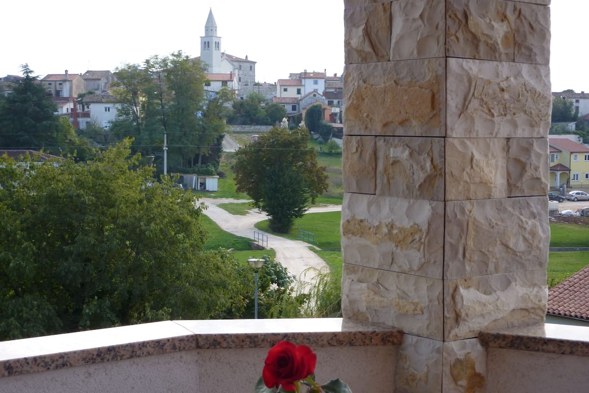 A stone pillar with a red rose in the foreground, a pathway leading to a town with buildings and a church in the background.