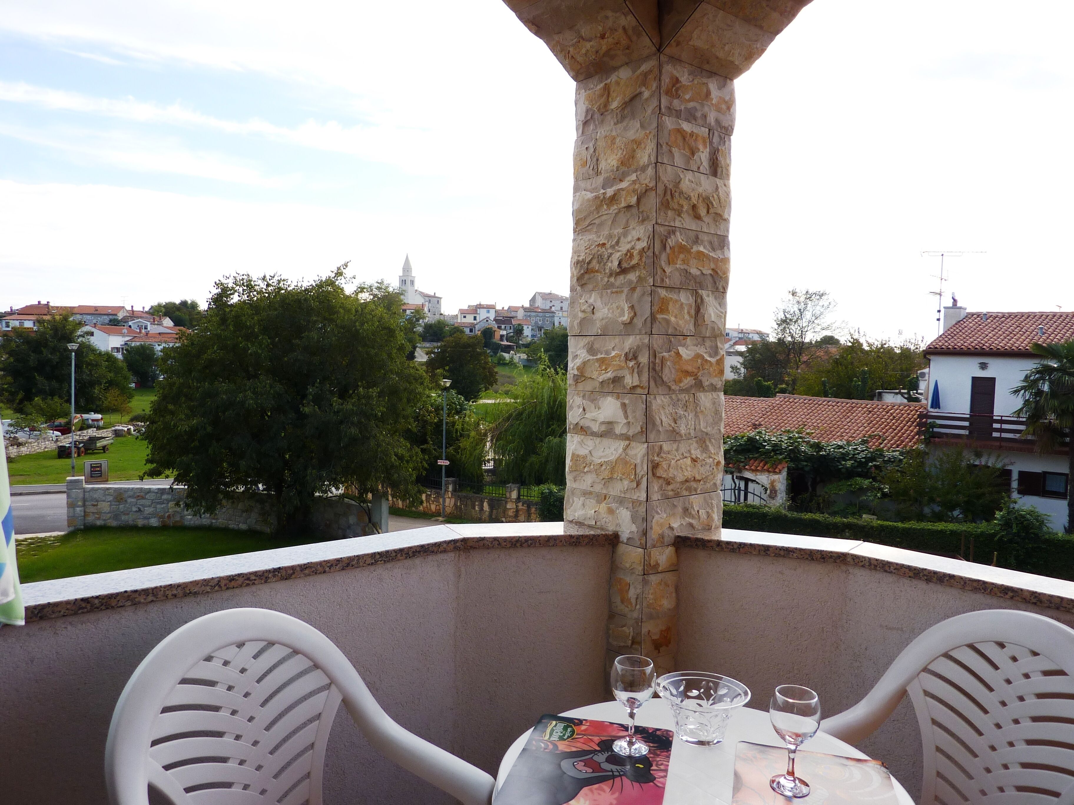 A balcony with a stone pillar, two white chairs, a table with a glass, and a view of a town with houses and a church.