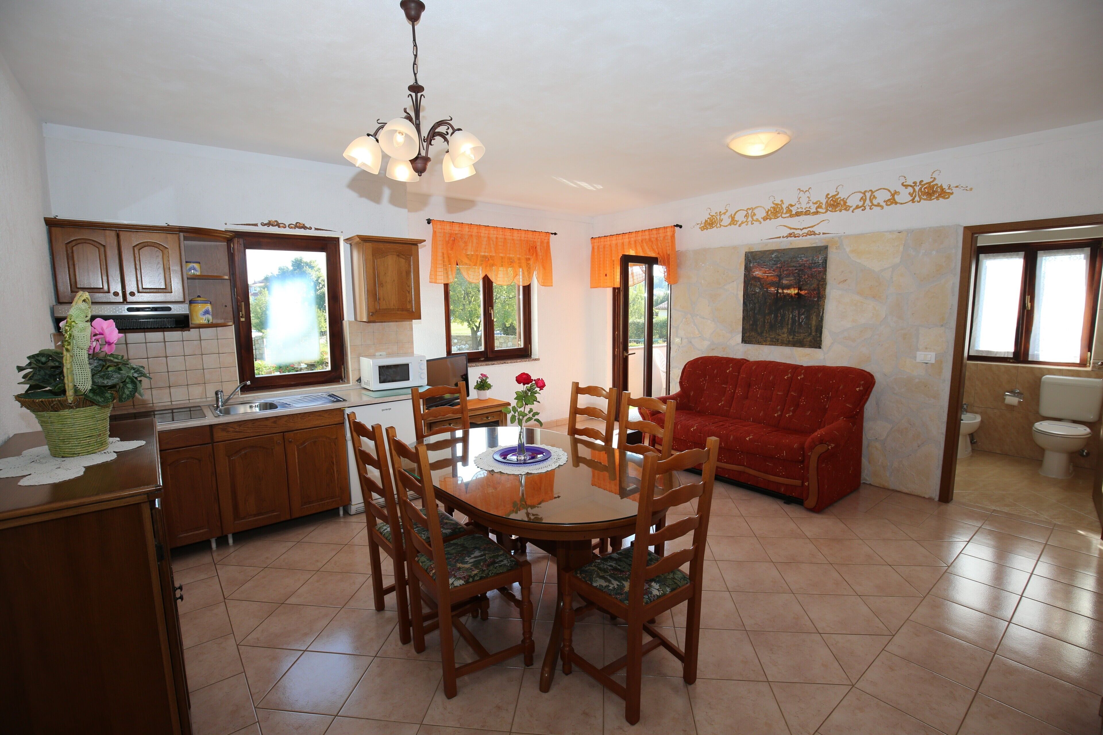 A dining area with a round table, chairs, a red sofa, and a kitchen in the background.