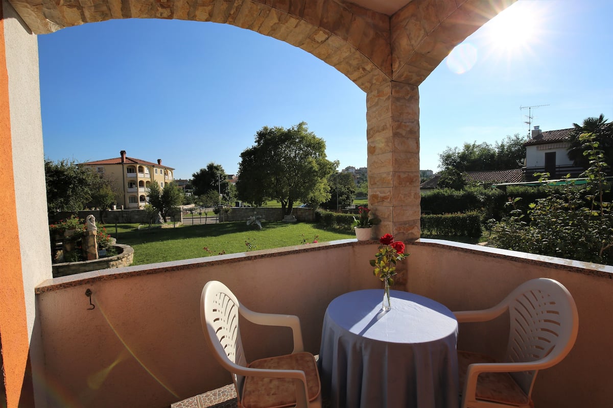 A balcony with a table and chairs, overlooking a garden and buildings.