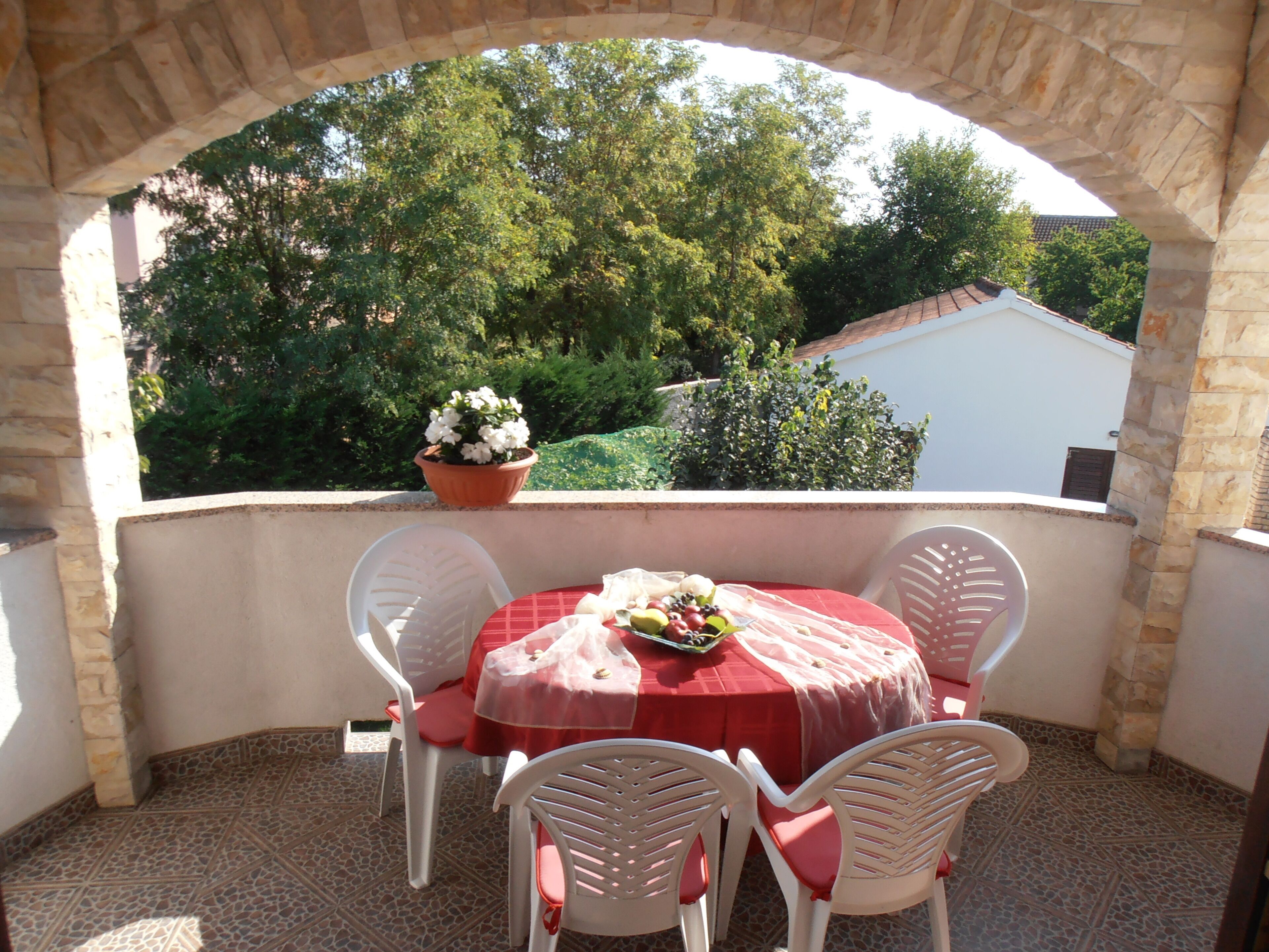 A covered outdoor patio with a round table and four chairs, a potted plant, and a view of trees and a building in the background.