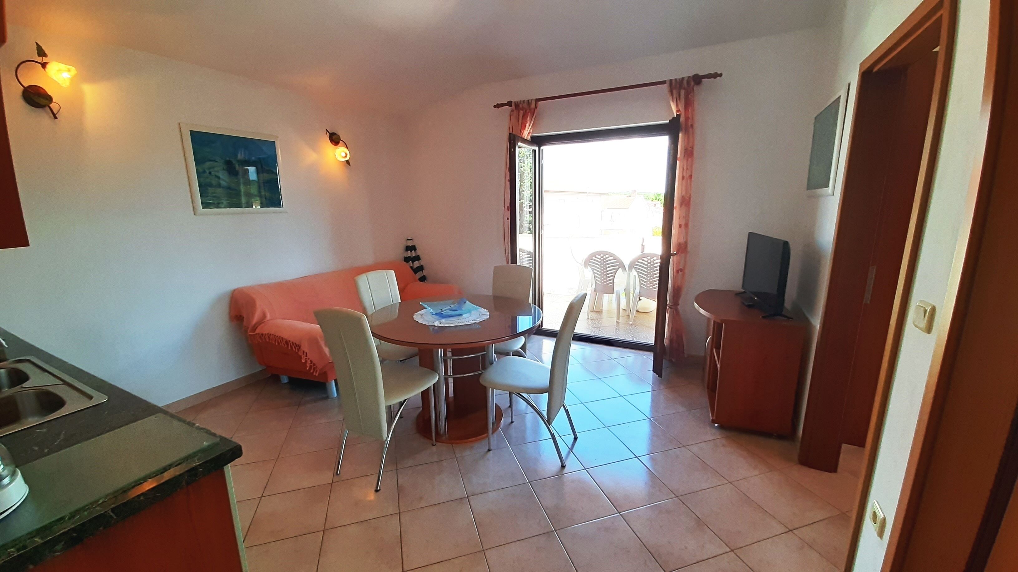 A compact room with a dining table, chairs, a sofa, a television, and a kitchen area.