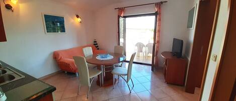 A compact room with a dining table, chairs, a sofa, a television, and a kitchen area.