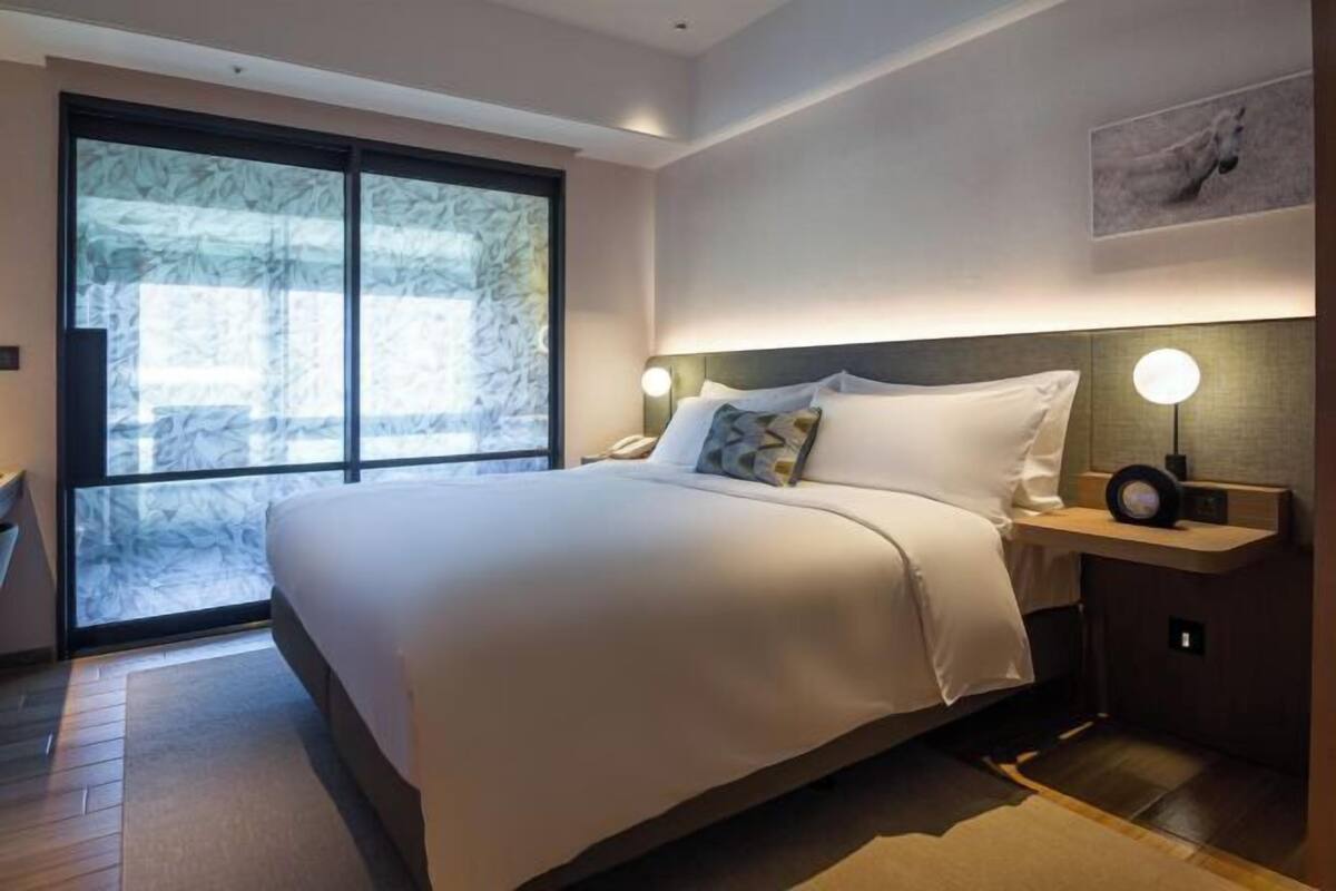 Een moderne hotelkamer met een groot bed, een nachtkastje met een lamp en uitzicht door een groot raam.
