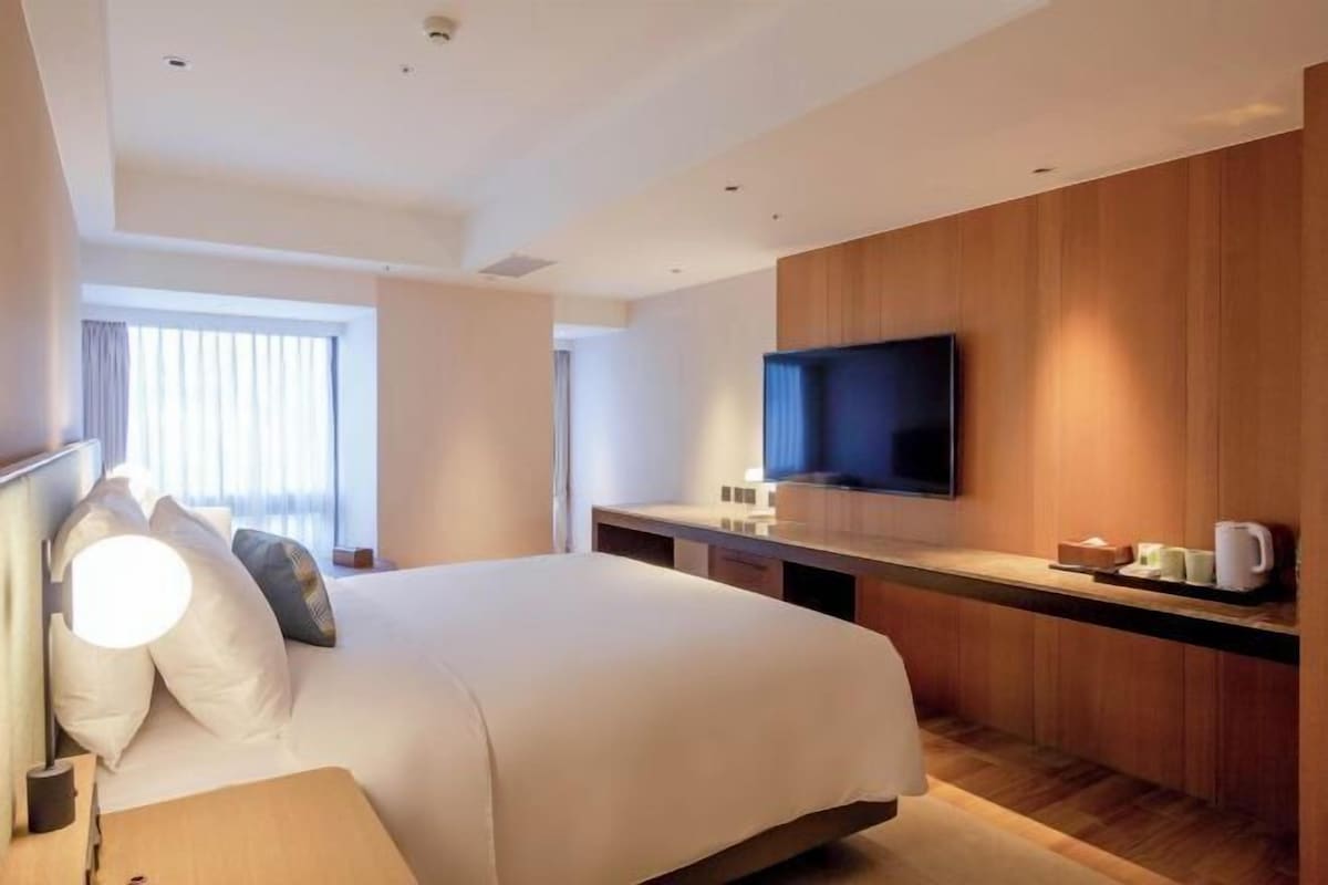 Una habitació d'hotel moderna amb un llit gran, un televisor de pantalla plana fixat a una paret de fusta, una taula de nit amb una làmpada i una finestra amb cortines.