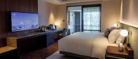 A modern hotel room with a large bed, a desk with a flat-screen TV, a seating area, and a view of the outdoors.