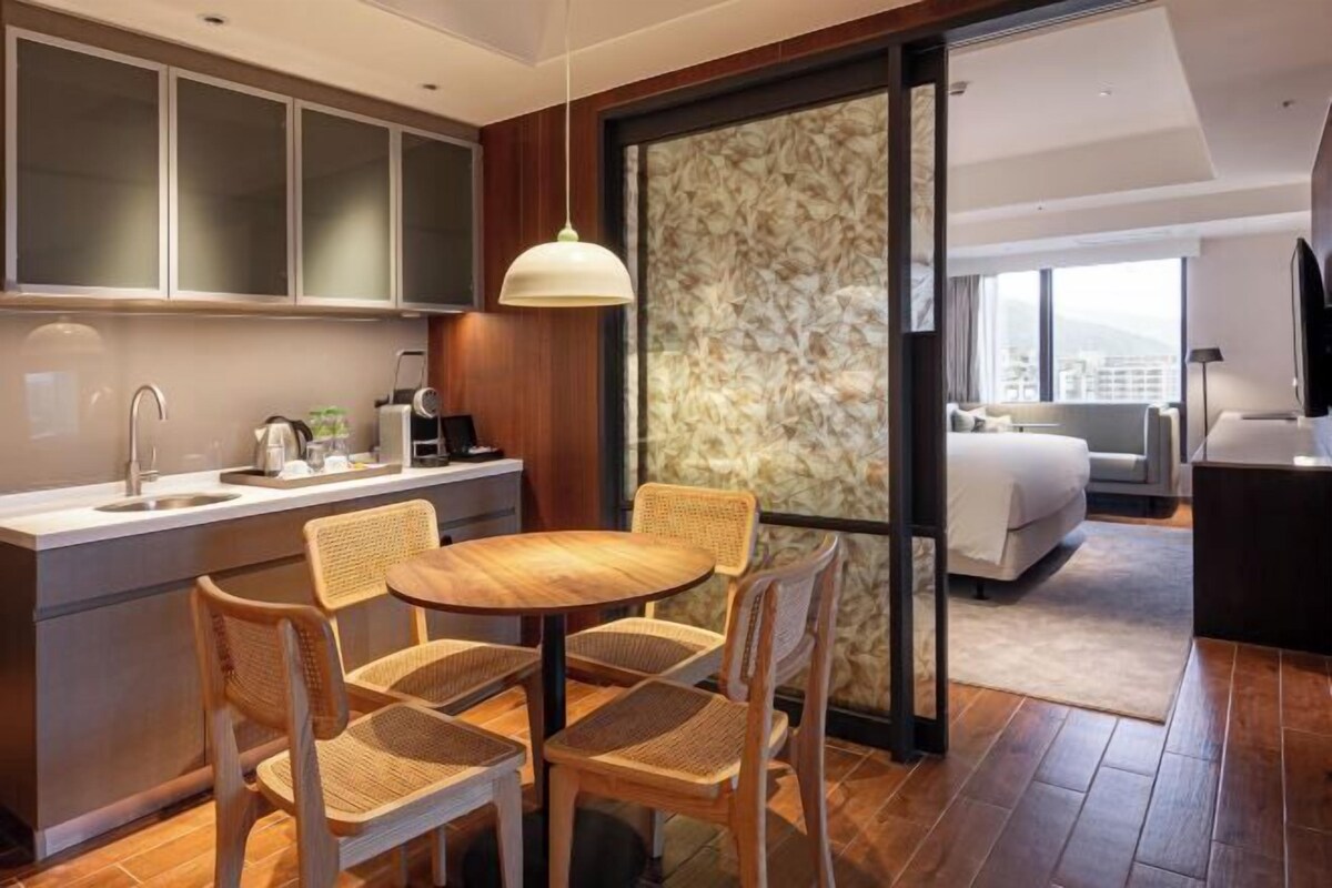 A modern hotel room with a kitchen area, a dining table with chairs, a large bed, and a TV.