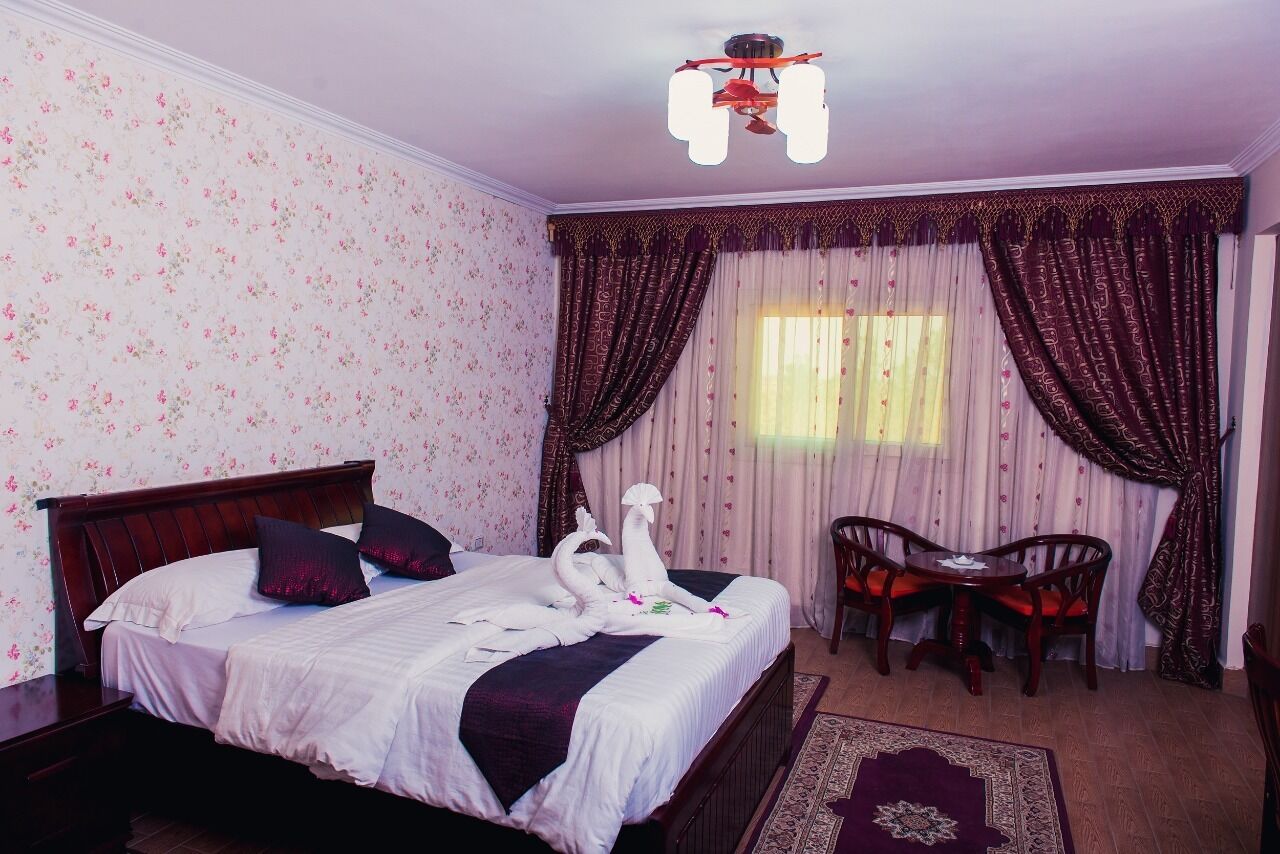 A neatly made bed with white linens and decorative swan-shaped pillows, a wooden headboard, a small round table with chairs, and a patterned rug.