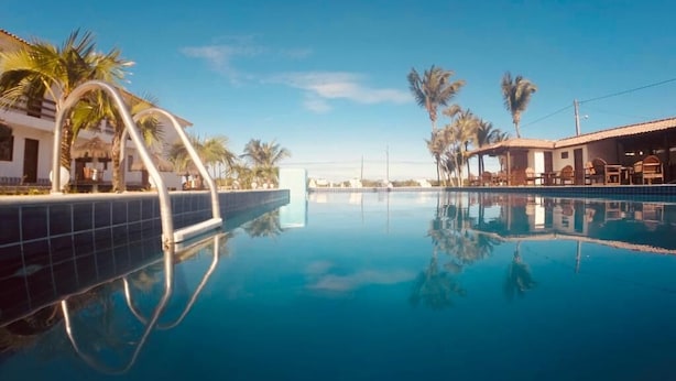 A clear blue pool with a tiled edge, surrounded by palm trees and a building with a tiled roof.