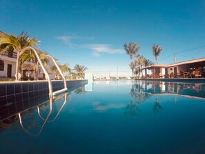 A clear blue pool with a tiled edge, surrounded by palm trees and a building with a tiled roof. - Porto Princesa Praia Hotel (Santa Cruz Cabralia)