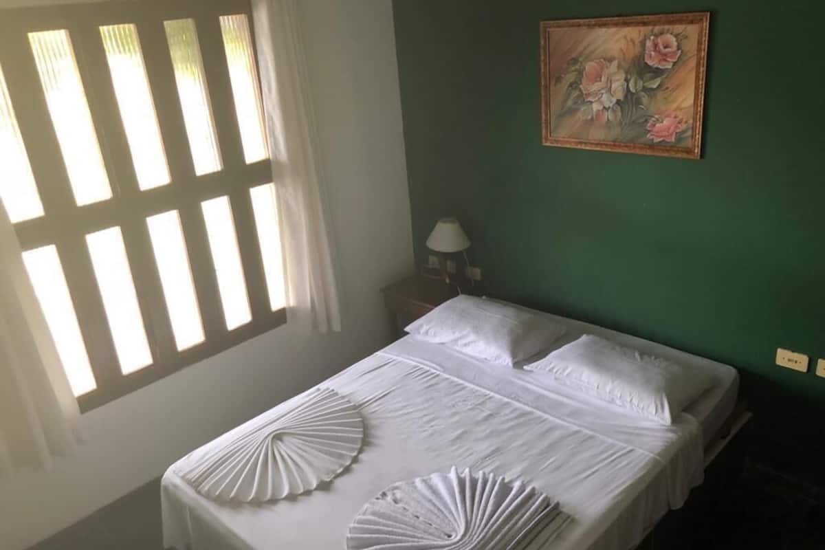 Ein ordentlich bezogenes Bett mit weißen Leinen und dekorativen Kissen, ein Gemälde an der Wand und ein Fenster mit Jalousien.