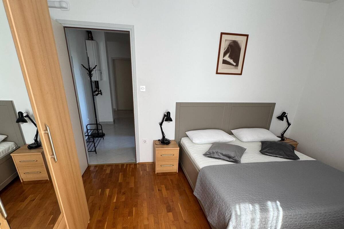 Un dormitorio con piso de madera, una cama con manta gris, dos mesitas de noche con lámparas, un armario y un cuadro en la pared.