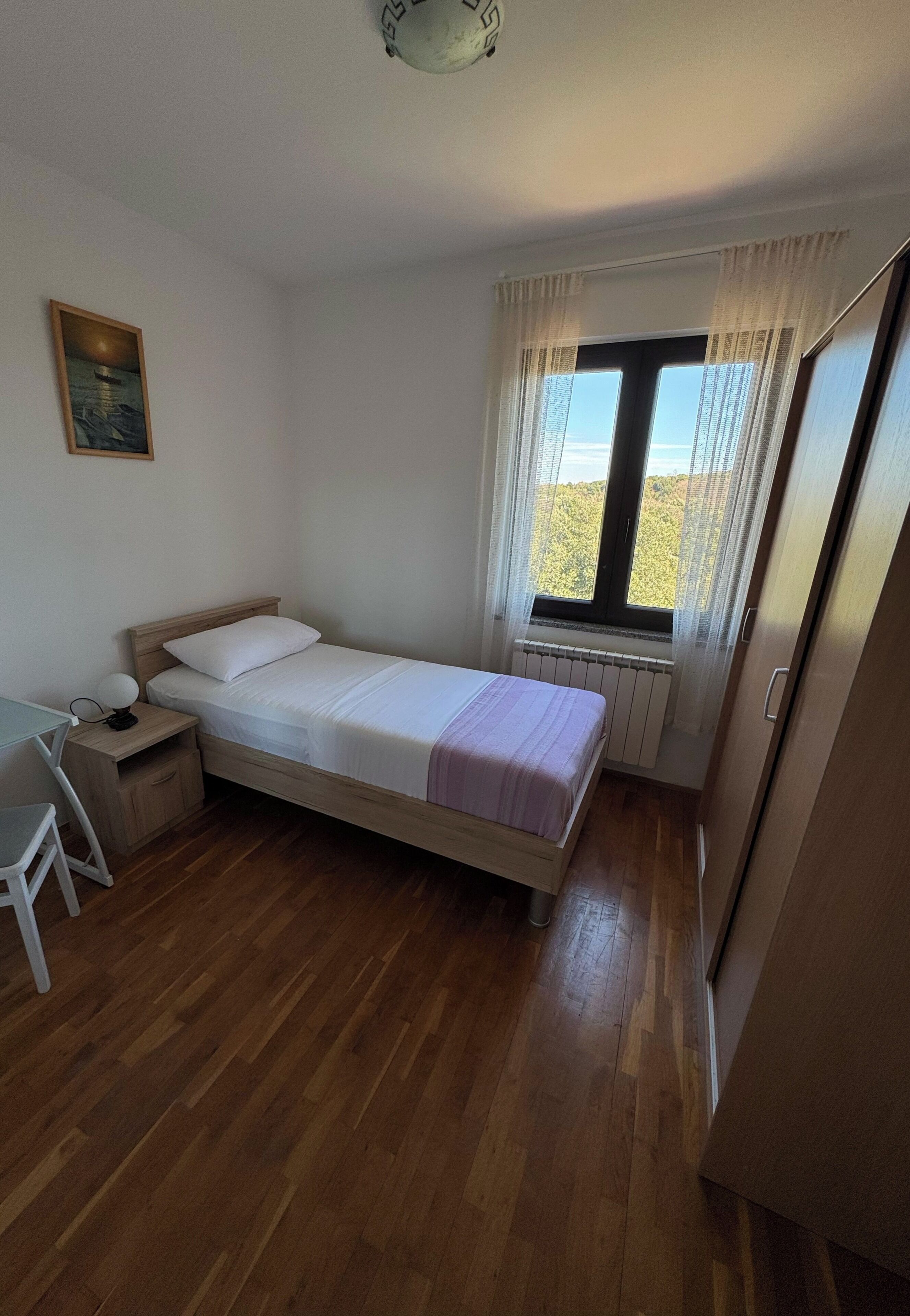 A bedroom with a bed, a desk, a chair, a radiator, a window with curtains, and a door.