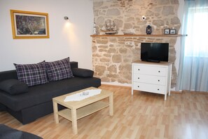 Una sala de estar con sofá, mesa de centro, televisor sobre un mueble, pared de piedra y estante con objetos decorativos.