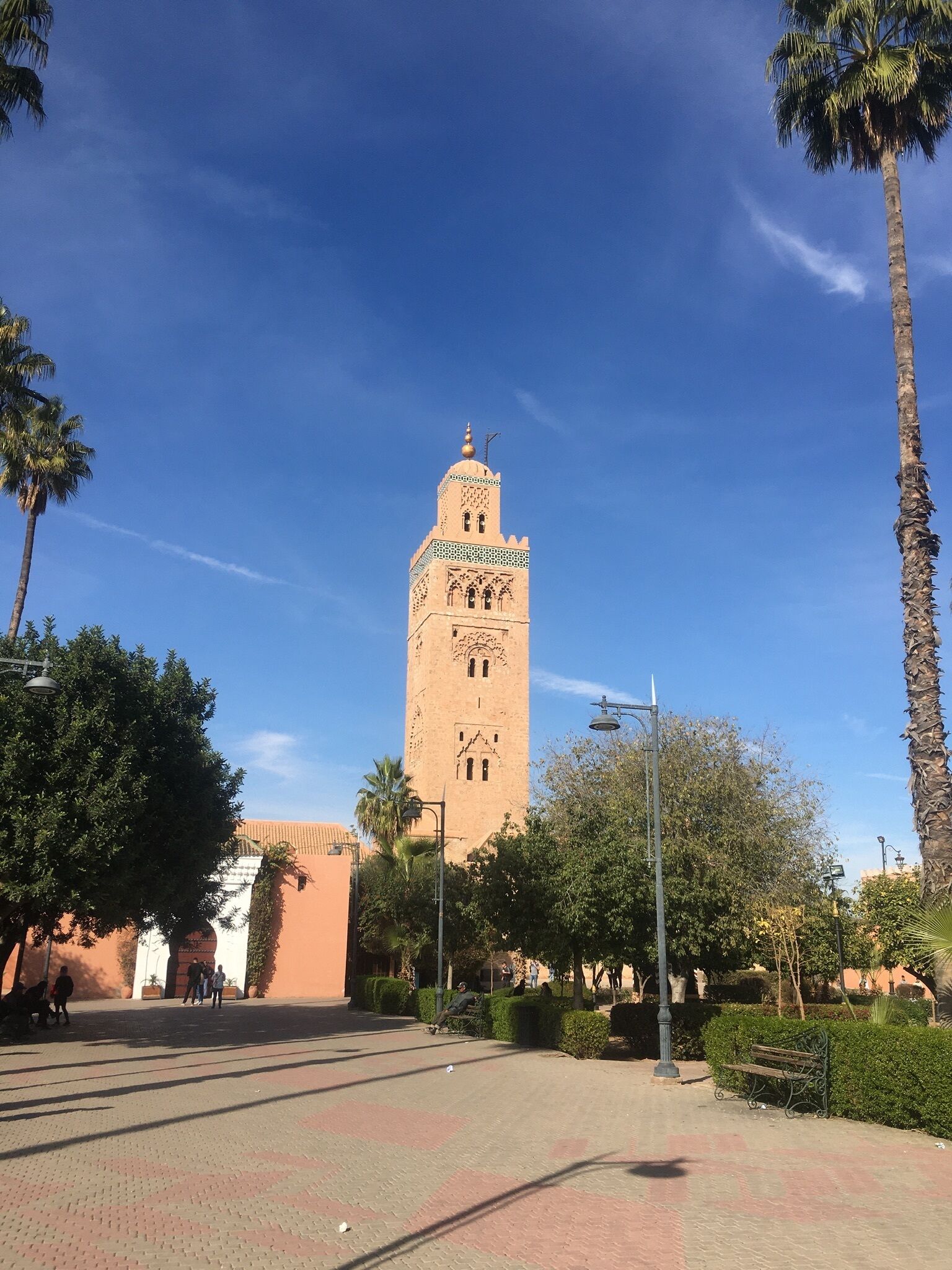 A tall tower with intricate designs and a dome on top, surrounded by palm trees and a paved walkway.