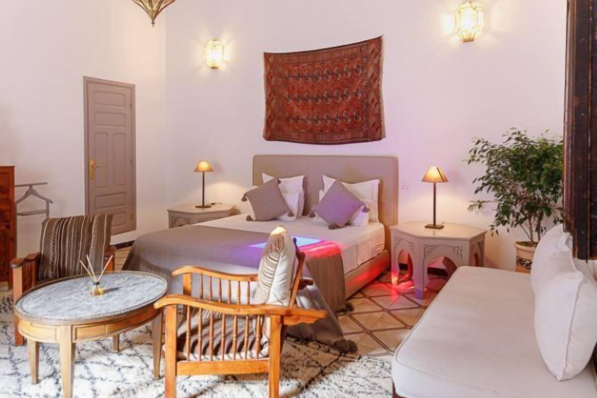 Un dormitorio con cama, mesitas de noche, una silla, un sofá, un tapiz colgado y una planta en maceta.