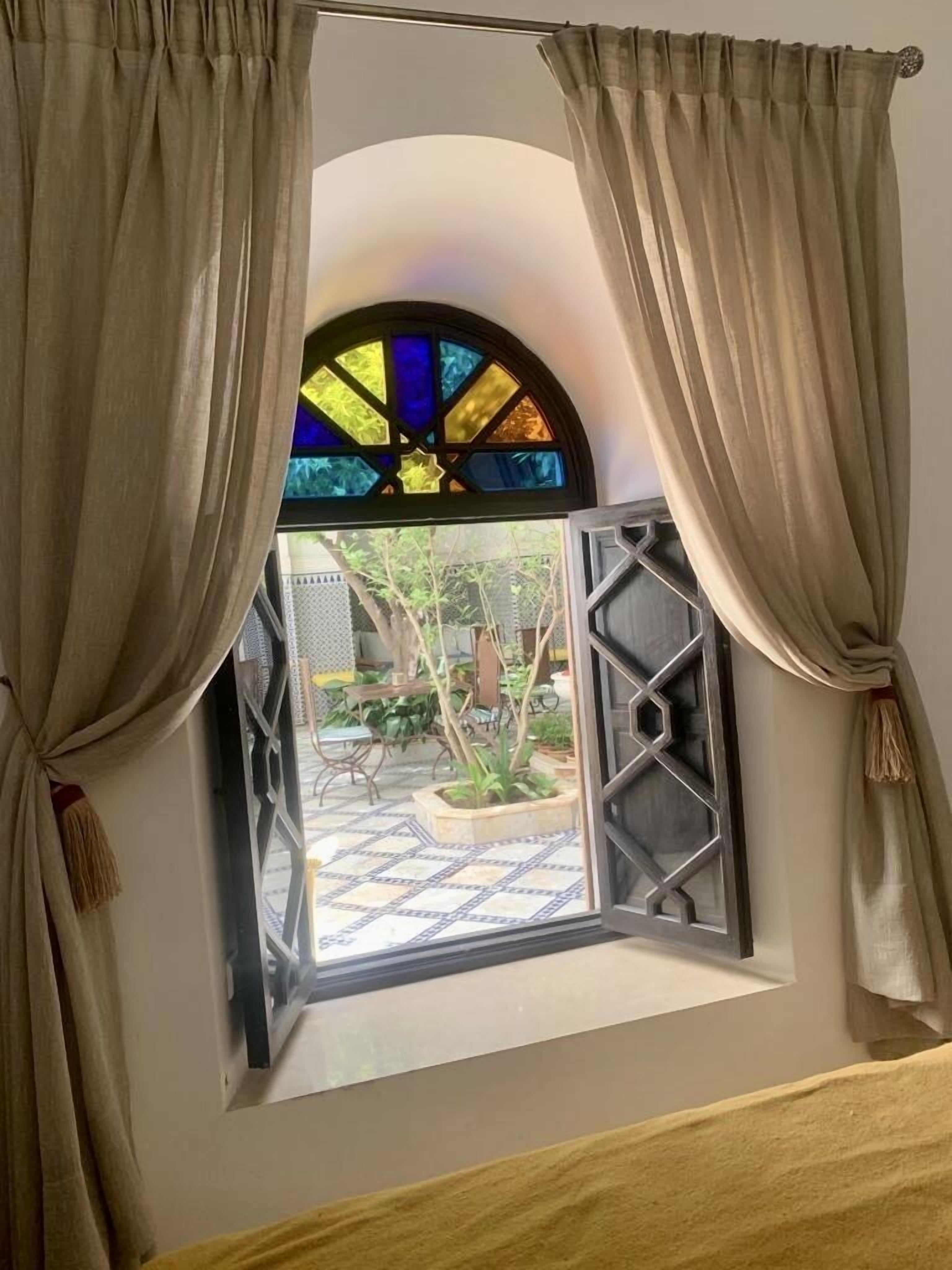 A window with beige curtains, a stained glass design, and an open view of an outdoor patio area with plants and furniture.