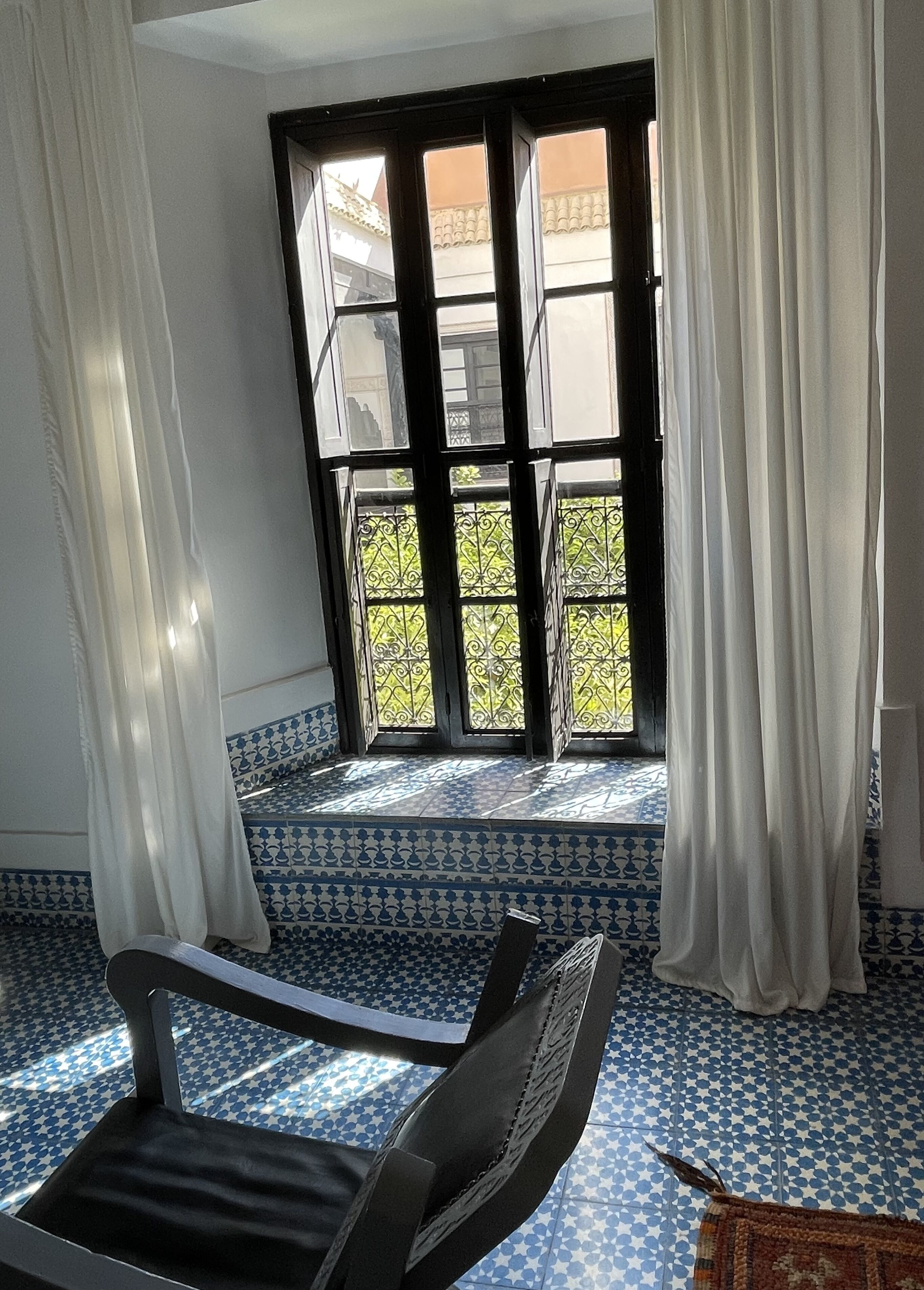 A room with a patterned blue and white floor, a black chair, a window with white curtains, and a view of a balcony with metal railings.