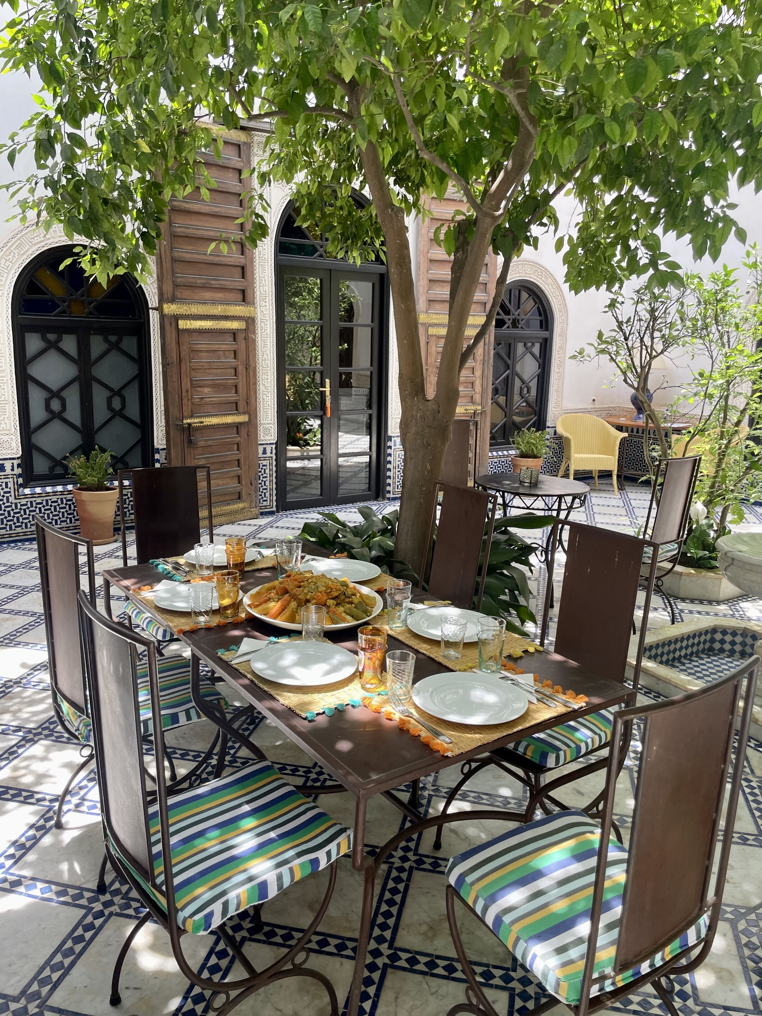 Une table à manger dressée avec des assiettes, des verres et divers plats, entourée de chaises à rayures et d’un arbre qui offre de l’ombre.